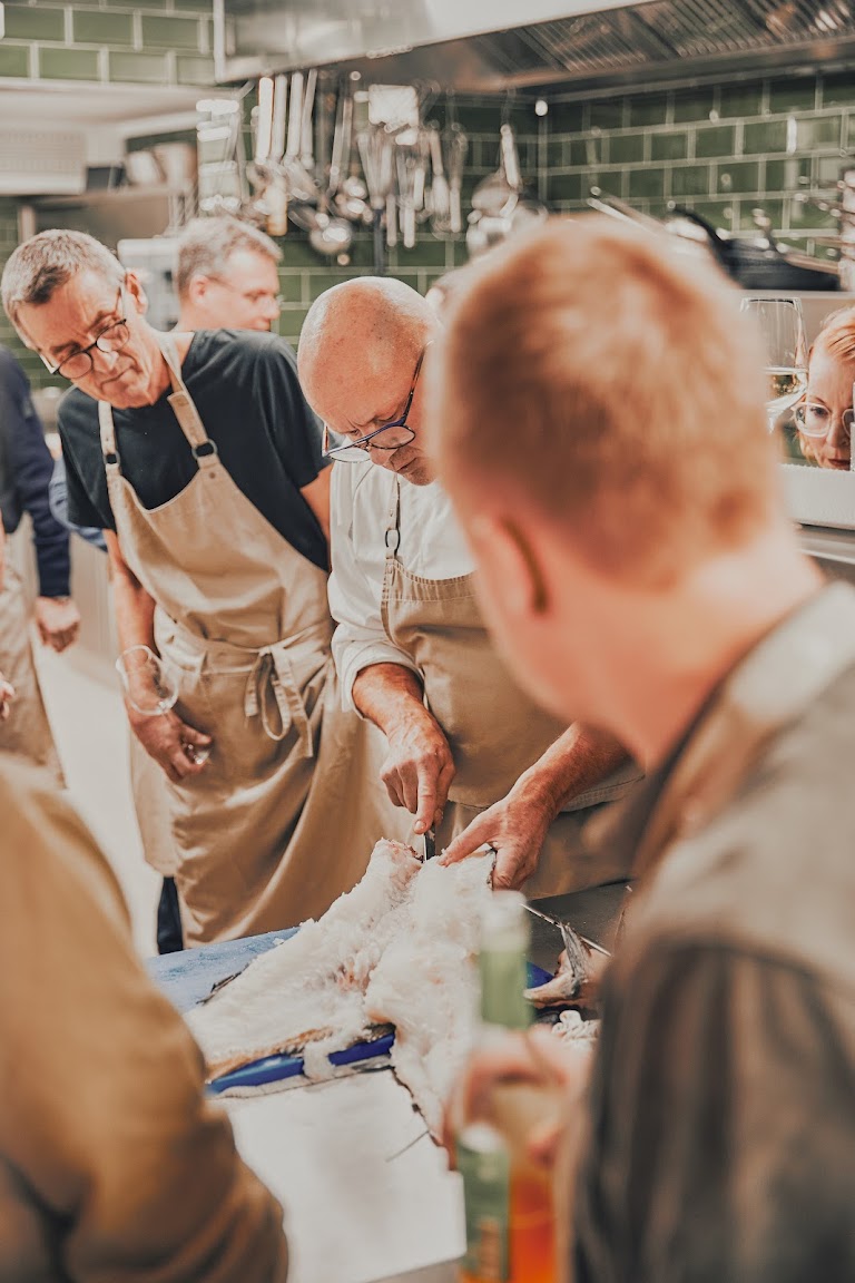 Rainer Trautenbach und Teilnehmer schneiden gemeinsam Fisch in der grün gekachelten Profiküche